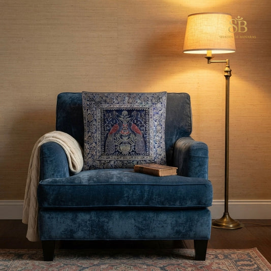 Blue brocade Banarasi cushion cover with peacock pair motif on a navy velvet armchair